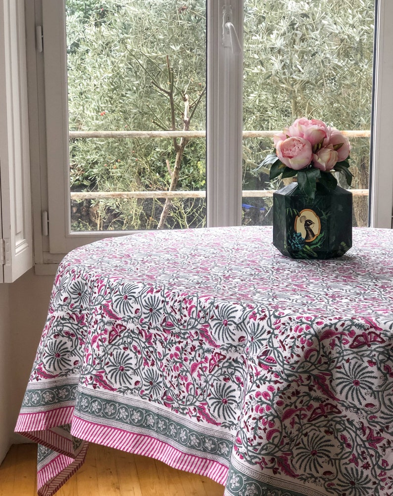 May include: A white tablecloth with a pink and gray floral pattern. The tablecloth has a pink and gray border. A vase with pink flowers sits on the tablecloth.