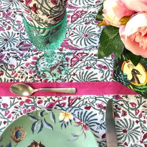 May include: A table setting with a green and white patterned tablecloth, a green glass, a white napkin, a green plate with a tiger design, a silver spoon, and a silver knife. The tablecloth has a pink border. There are pink flowers in a vase in the background.