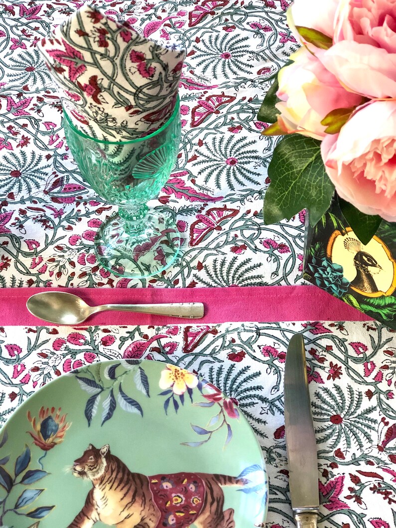 May include: A table setting with a green and white patterned tablecloth, a green glass, a white napkin, a green plate with a tiger design, a silver spoon, and a silver knife. The tablecloth has a pink border. There are pink flowers in a vase in the background.
