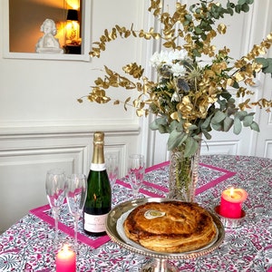 May include: A festive table setting with a pink and white patterned tablecloth, a golden-leafed floral arrangement, a bottle of champagne, two champagne flutes, a round pie on a silver stand, and pink candles.