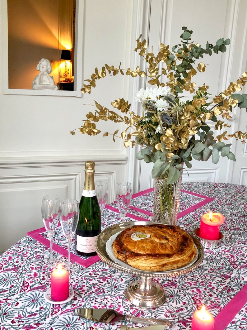 May include: A festive table setting with a pink and white patterned tablecloth, a golden-leafed floral arrangement, a bottle of champagne, two champagne flutes, a round pie on a silver stand, and pink candles.