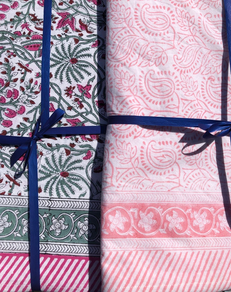 May include: Two fabric squares tied together with blue ribbon. One square has a white background with pink floral patterns. The other square has a white background with green and pink floral patterns.