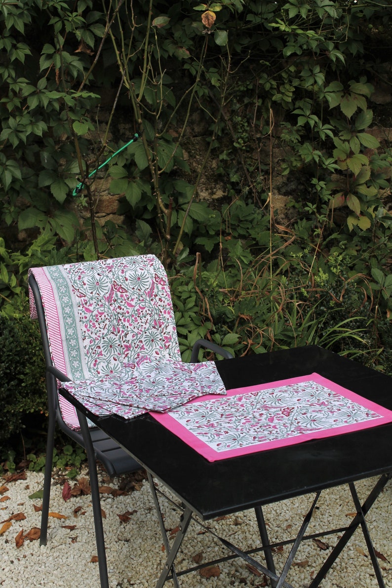 May include: A black metal table with a pink and white floral patterned tablecloth and matching placemats. The table is set up outdoors with a chair in the background.