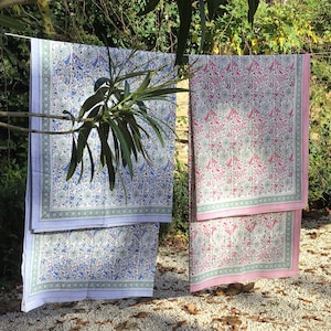 May include: Two patterned cotton tablecloths hanging on a clothesline outdoors. The tablecloth on the left is white with blue and green floral designs. The tablecloth on the right is white with pink and green floral designs.