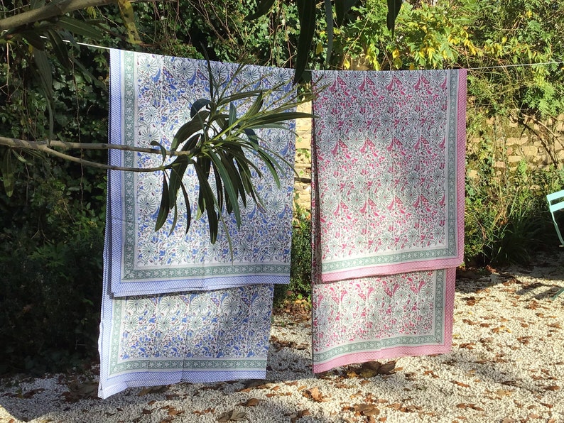 May include: Two patterned cotton tablecloths hanging on a clothesline outdoors. The tablecloth on the left is white with blue and green floral designs. The tablecloth on the right is white with pink and green floral designs.