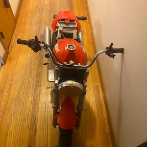 May include: A red and silver toy motorcycle with black handlebars and a black tyre. The motorcycle has a red fuel tank and a headlight. The word "Ducati" is visible on the fuel tank.