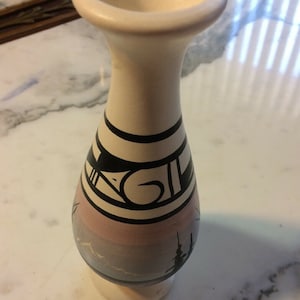 Peut inclure: Un vase en céramique de couleur crème avec un dessus évasé et un col étroit. Le vase présente un motif géométrique noir et blanc au milieu, avec une scène de paysage dans les tons de rose, de gris et de bleu à la base. Le vase mesure environ 15 cm de haut.