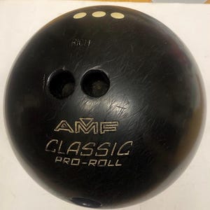 Puede incluir: Una bola de bolos negra con dos agujeros para los dedos y tres agujeros para el pulgar. La bola tiene el texto "AMF CLASSIC PRO-ROLL" en letras plateadas. La palabra "RICH" también es visible en la bola.