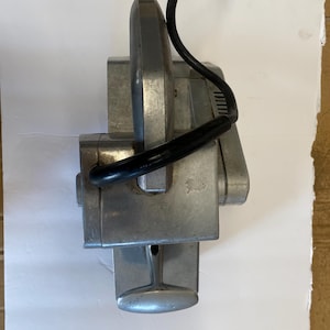 May include: A vintage, silver-colored metal tool with a black handle and power cord. The tool has a rectangular body with a curved top and a lever-like handle. The power cord is black and coiled.