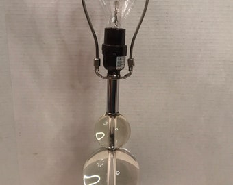 Könnte beinhalten: Eine Tischlampe aus klarem Glas und Chrom. Die Lampe hat einen quadratischen Sockel, einen Chromschaft und zwei klare Glaskugeln. Die Lampe hat einen Chrom-Lampenschirmhalter und eine Glühbirne. Die Lampe ist eine dekorative Leuchte.