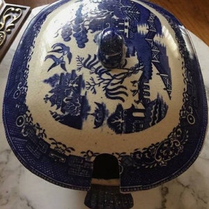 May include: A blue and white ceramic serving dish lid with a detailed Willow pattern design. The lid features a central knob and a scalloped edge. The design includes scenes of buildings, trees, and other elements.