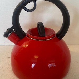 May include: A red, round tea kettle with a black handle and spout. The kettle has a glossy finish and a small, black handle on the lid. The large handle arches over the top of the kettle.