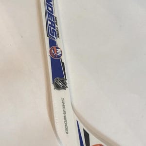 May include: White and blue hockey stick with the words "ISLANDERS NEW YORK" in blue. The stick has the NHL logo and the team's logo. The brand name "SHERWOOD" is also visible.