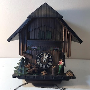 May include: A dark brown wooden cuckoo clock with a pitched roof, featuring a clock face with Roman numerals. The clock has decorative elements including a miniature figure, a tree, and a woodpile. The clock is hanging by a chain.