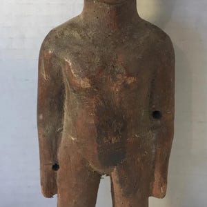 May include: A carved wooden figure with a dark brown finish. The figure is standing, with arms at its sides, and has a simple, stylized form. The wood shows signs of age and wear, adding to its rustic charm.