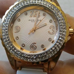 Può includere: Orologio da polso Akridoos color oro con quadrante bianco e lunetta incastonata con piccoli cristalli trasparenti. Il quadrante presenta lancette e indici dorati, con i numeri 3, 6 e 9 visibili. Il cinturino sembra essere bianco.