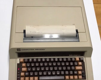 May include: A vintage beige Computer Devices Miniterm terminal with a full keyboard and a paper output slot. The keyboard has brown keys with white and black lettering. The terminal has a small paper printout.