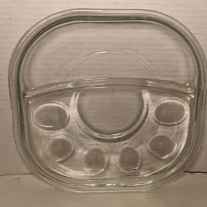 May include: Clear glass serving dish with a handle and multiple compartments. The dish has a rounded rectangular shape with a handle on one side and a central circular area surrounded by smaller oval indentations.