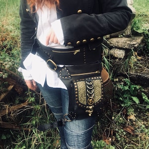 Könnte beinhalten: Ein schwarzer Ledergürtel mit einer großen, dekorativen Tasche im Schlangenhautmuster. Die Tasche hat goldfarbene Akzente und Schnallen. Der Gürtel wird über einem weißen Hemd und einer blauen Jeans getragen.