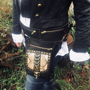 Könnte beinhalten: Ein schwarzer Leder-Steampunk-Gürtel mit einer Schlangenhaut-Tasche und goldfarbenen Beschlägen. Der Gürtel hat mehrere Schnallen und Riemen, und die Tasche hat einen Reißverschluss.