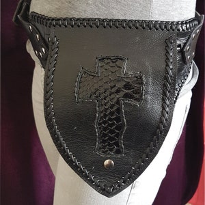 Może przedstawiać: Czarny skórzany pasek z dużym, tłoczonym wzorem krzyża. Pasek ma pleciony brzeg i jest ozdobiony srebrnymi ćwiekami.