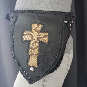 Może przedstawiać: Czarny skórzany pasek z wzorem krzyża ze skóry węża. Pasek ma pleciony brzeg i srebrną klamrę.