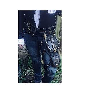 Könnte beinhalten: Ein schwarzer Ledergürtel mit goldenen Akzenten und eine passende schwarze Leder-Oberschenkel-Tasche mit Schlangenhautmuster. Der Gürtel hat eine Schnalle und die Tasche einen Reißverschluss.