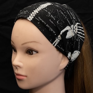 May include: Black headband featuring a white anatomical illustration of the human skeleton, including the spine, humerus, clavicle, ulna, and metacarpals. The headband has a black background with white stars.