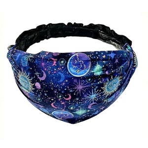 May include: A blue headband with a celestial print featuring moons, stars, and suns. The headband is made of a soft, stretchy fabric and has a black elastic band.