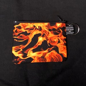 Puede incluir: Una bolsa negra con cremallera con un diseño de llamas. La bolsa tiene un llavero plateado. El texto de la etiqueta dice "Been England Hand Made By"