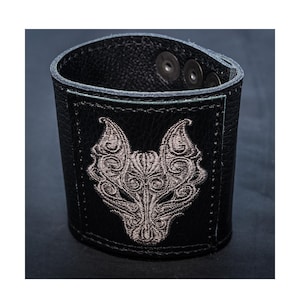 May include: Black leather bracelet with a detailed embroidered wolf design in gray. The bracelet has three silver snaps.