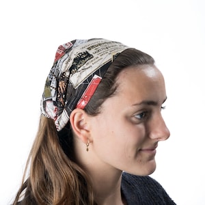 Puede incluir: Una mujer con el pelo largo y castaño lleva una diadema negra y roja con un estampado vintage de tren y texto. La diadema está anudada en la parte posterior de la cabeza.