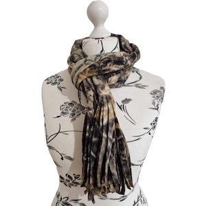 Puede incluir: Una bufanda de franela marrón y beige con un patrón de camuflaje y flecos. La bufanda se muestra en un maniquí blanco con un patrón floral.