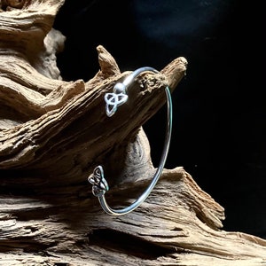 May include: A silver bangle bracelet with a Celtic knot design on each end. The bracelet is displayed on a piece of driftwood.
