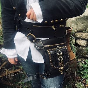 Könnte beinhalten: Schwarze Steampunk-Hüfttasche aus Leder mit mehreren Fächern, Reißverschlüssen und messingfarbener Hardware. Die Tasche hat eine dekorative Platte mit strukturiertem Muster und einem zahnradförmigen Schmuck. Getragen mit einer schwarzen Jacke und Jeans.