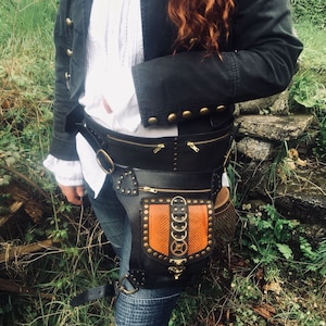 Könnte beinhalten: Ein schwarzer Ledergürtel im Steampunk-Stil mit einer großen, verzierten Metallschnalle und einer kleinen, orangefarbenen Ledertasche mit Reißverschluss.