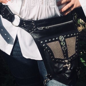 Könnte beinhalten: Eine schwarze Leder-Bauchtasche mit Reißverschluss, silbernen Nieten und einem braunen Lederpanel mit Zahnrädern. Die Bauchtasche wird an der Taille getragen.