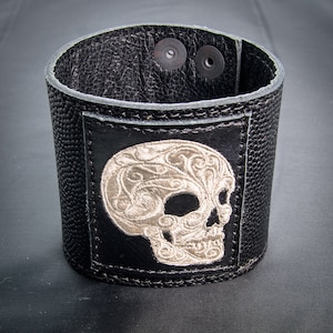 Peut inclure: Bracelet en cuir noir avec un motif de crâne brodé argenté. Le crâne présente des détails complexes et un motif floral.