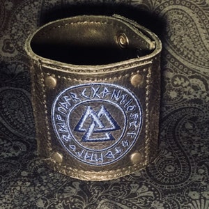 Puede incluir: Un brazalete de cuero marrón con un círculo bordado en azul que contiene un símbolo de triángulo triple y el texto "RAIDO URNIR", una inscripción rúnica nórdica.