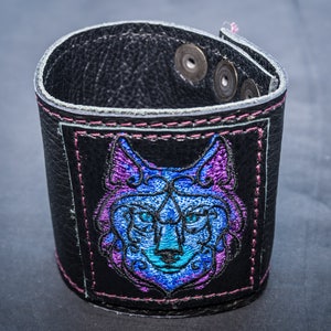 Peut inclure: Un bracelet en cuir noir avec une bordure cousue rose et un motif de tête de loup brodé bleu et violet.