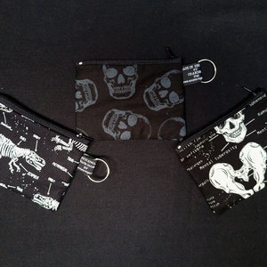 May include: Three black zippered pouches with white anatomical illustrations. The pouches feature a dinosaur skeleton, a human skull, and a human pelvis.