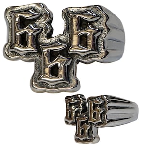 Puede incluir: Anillo plateado con las letras "RBG" en fuente gótica. El anillo tiene una banda gruesa con ranuras verticales. Las letras están en relieve y tienen una apariencia detallada y texturizada.