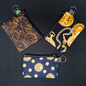 Pode incluir: Três pequenas bolsas com zíper com designs diferentes. A primeira bolsa tem um fundo preto com mostradores de relógio dourados. A segunda bolsa tem um fundo preto com relógios de bolso dourados e correntes. A terceira bolsa tem um fundo azul escuro com sóis dourados.