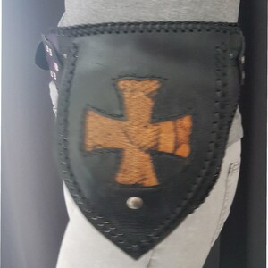 Könnte beinhalten: Ein schwarzer Ledergürtel mit einem großen, braunen Kreuzdesign. Der Gürtel wird um die Taille getragen und hat ein dekoratives Nähtdetail.