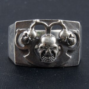 May include: A silver ring with a design of three skulls. The skulls are detailed and have a menacing look.