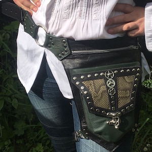 Könnte beinhalten: Ein schwarzer Ledergürtel mit einer grünen Ledertasche und silbernen Akzenten. Die Tasche hat einen Reißverschluss und einen dekorativen Metallverschluss. Der Gürtel hat eine silberne Schnalle und einen silbernen Ring.