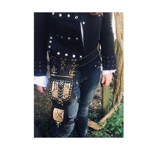 Könnte beinhalten: Ein schwarzer Ledergürtel mit einem Oberschenkelholster aus Schlangenleder und mehreren Metallverzierungen. Der Gürtel hat eine Schnalle und wird über einer schwarzen Jacke mit silbernen Nieten getragen.