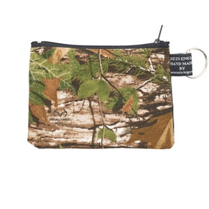 May include: A small, rectangular camouflage zippered pouch with a black zipper and a silver key ring. The pouch features a Realtree logo and a tag that reads "MADE IN ENGLAND HAND MADE BY oceania-legend". The pattern includes shades of green, brown, and tan.