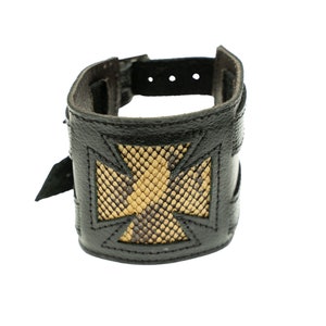 Peut inclure: Manchette de poignet en cuir noir avec un motif de croix décorative. La croix est remplie d'un motif peau de serpent dans des tons de beige et de marron. La manchette a une fermeture à boucle.