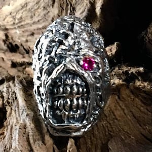 May include: A silver ring with a detailed, grotesque face design. The face has a large, open mouth with teeth and a single pink gemstone eye.